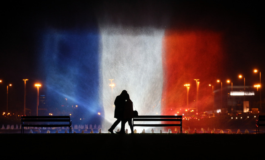 A young couple stands in front of illuminated waters with the colors of the French national flag on November 16, 2015 to pay tribute to victims of the attacks claimed by Islamic State which killed at least 129 people and left more than 350 injured on November 13. AFP PHOTO / STRINGER / AFP / STR